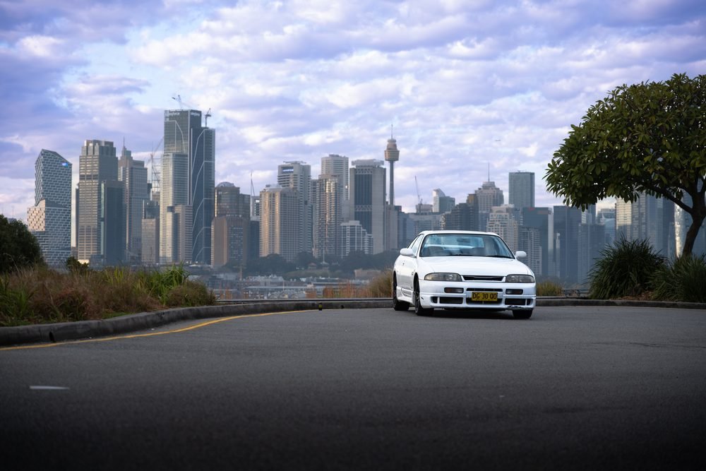 White Nissan Skyline GTS centred on a road with the Sydney CBD skyline behind it at sunset
